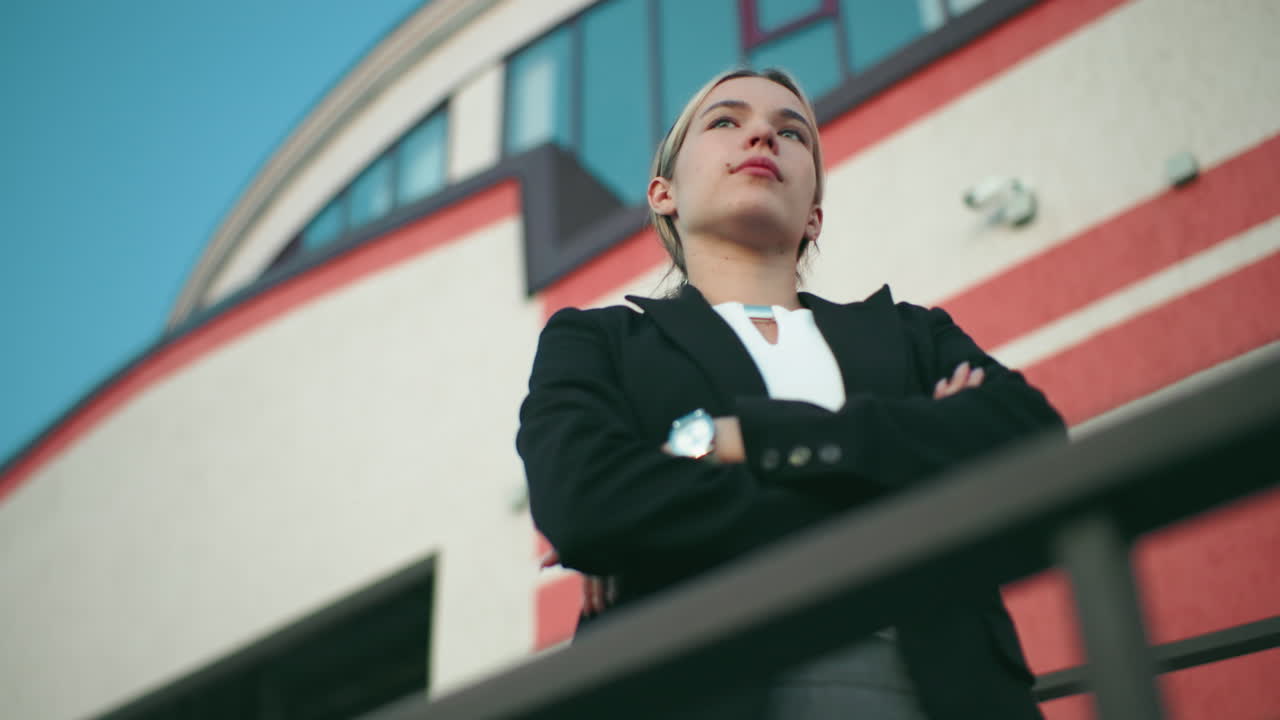 Woman in black blazer and white top stands confidently with arms folded, gazing into distant sky in front of modern building, exuding ambition
