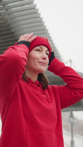 mujer de negocios con capucha roja ajusta su gorra mientras sonríe y mira a la distancia, el fondo incluye dosel moderno, colina nevada y postes de luz en un clima de invierno nebuloso
