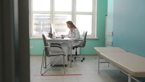 doctora sonriente escribiendo en papel mientras se sienta en el escritorio en su sala de consulta