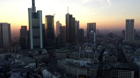 AERIAL:-View-of-Frankfurt-am-Main,-Germany-Skyline-with-sunflair-between-skyscrapers-in-Beautiful-Sunset-Sunlight-in-Winter-Haze