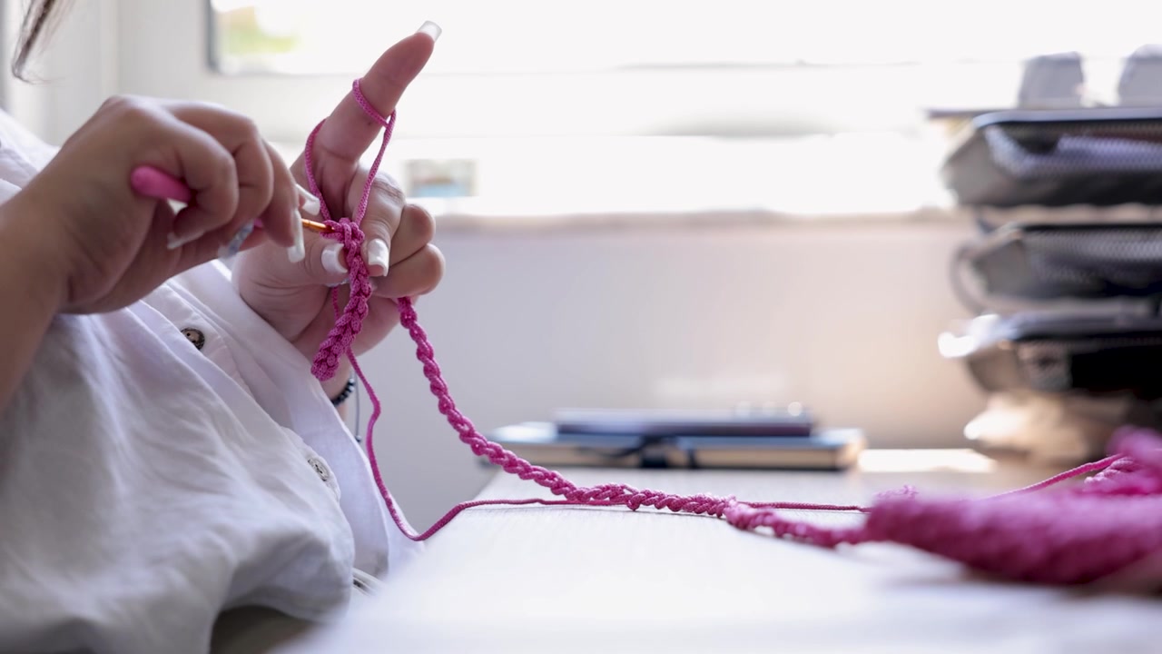 Close-up of a woman crocheting with bright pink yarn and a crochet hook at a worktable. Handmade textile craft showing traditional handwork, creativity and artisan production.