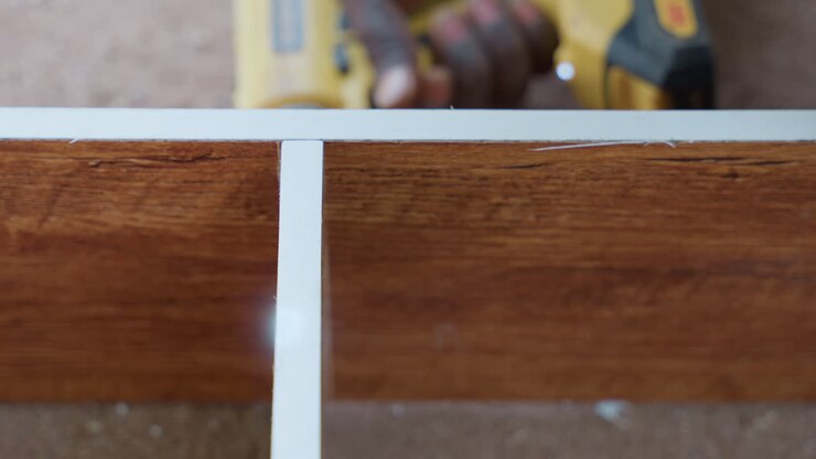 Primo piano dell'installazione di una modanatura su legno tinto, bordo bianco allineato in una scanalatura con mani ferme, trapano appoggiato sullo sfondo, venatura della superficie e linee pulite visibili, finitura precisa per la falegnameria.