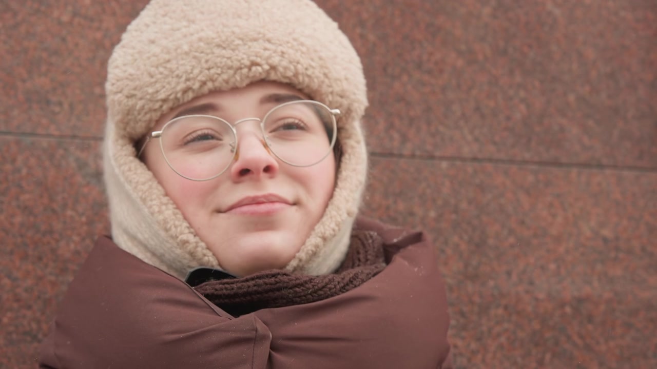 Close-up motion sequence of person wearing fuzzy beige hat and round metal eyeglasses raising hand to adjust glasses and hat, squinting upward then lowering eyes into relaxed closed-eyes smile while