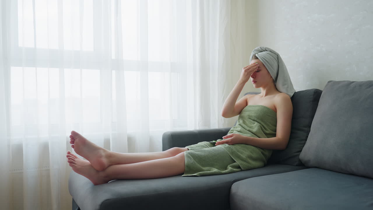 Lady sitting on couch wrapped in towel and towel turban, gently massaging forehead with cucumber slice as part of calming skincare routine in soft daylight from window in peaceful indoor setting