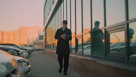 magnate de los negocios en abrigo negro caminando cerca de un edificio de vidrio, mirando a los coches estacionados, luego centrándose de nuevo en la tableta que se refleja en el vidrio, las características de fondo edificio borroso en la lejanía