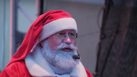 a positive old american santa claus speaks to the audience into microphone while standing on stage a positive old american santa claus speaks to the audience into a microphone while standing on stage new years santa claus wishes people a merry christmas makes emotional gestures new year party