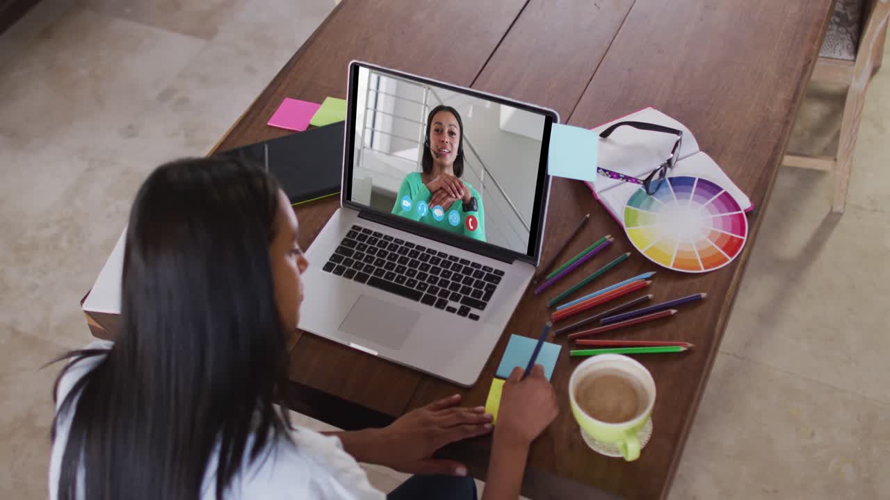 Mujer caucásica usando una laptop en videollamada con una colega, tomando notas