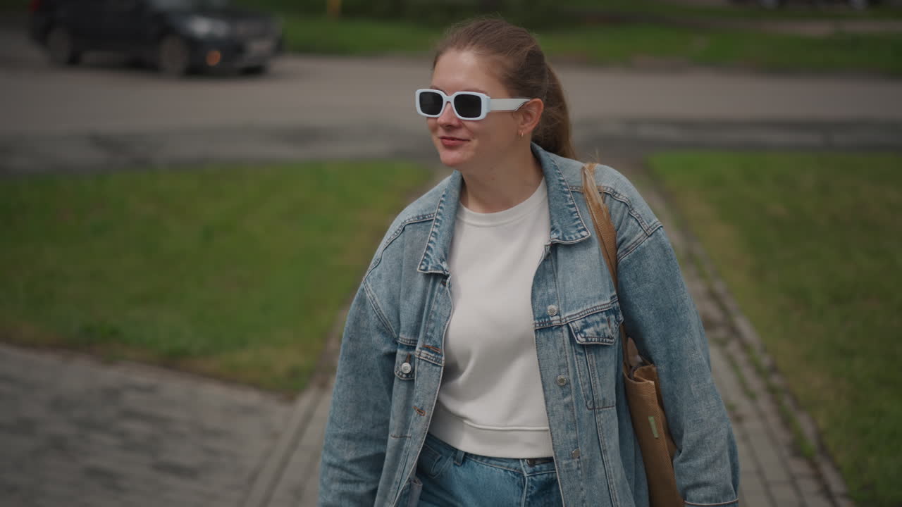 Young woman walking sidewalk denim jacket, sunglasses, tote bag, soft overcast light, shallow depth, intimate close framing, reflective expression, park path, cinematic color grading, casual fashion