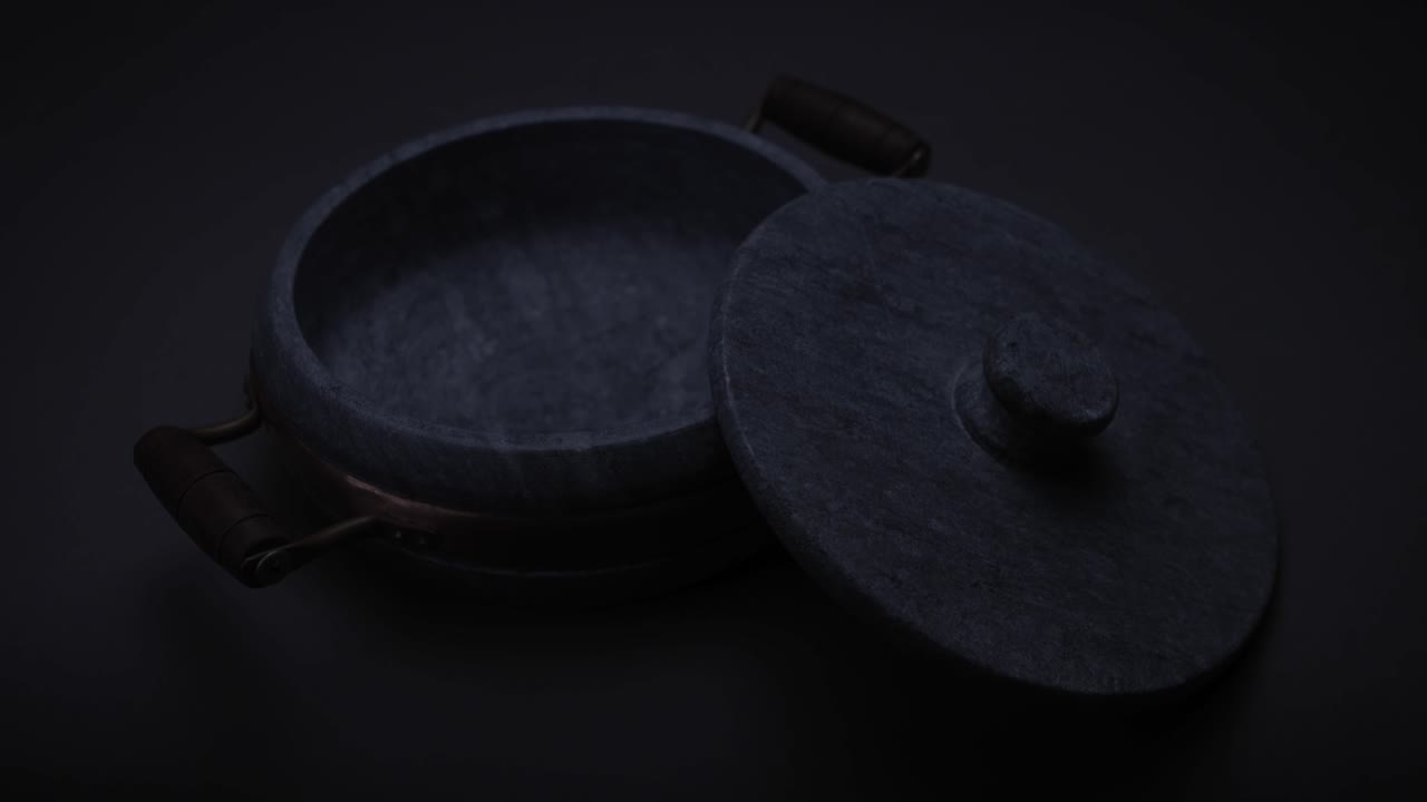 Smooth round heavy soap stone casserole with brass element and wooden handle with the lid resting half on it on a grey seamless surface with the light changing to cast moving shadows