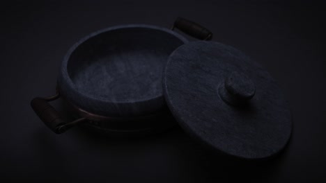 smooth round heavy soap stone casserole with brass element and wooden handle with the lid resting half on it on a grey seamless surface with the light changing to cast moving shadows