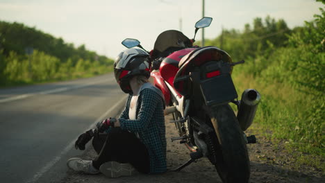 a female biker, wearing a helmet, sits cross-legged on the ground next to her red power bike parked by a rural road, she appears to be calmly observing her surroundings