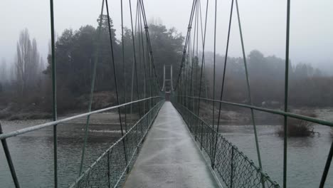 Drohne,-Die-Auf-Einem-Hängenden-Gehweg-über-Den-Fluss-Fliegt,-Mit-Nebligem-Wald-In-Der-Ferne---Drohnenansicht