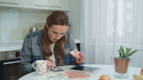 mujer sonriente hablando teléfono inteligente durante el desayuno en la cocina moderna.