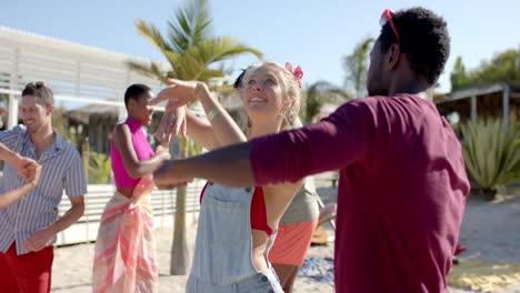 happy diverse group of friends dancing on beach with beach house and palm trees