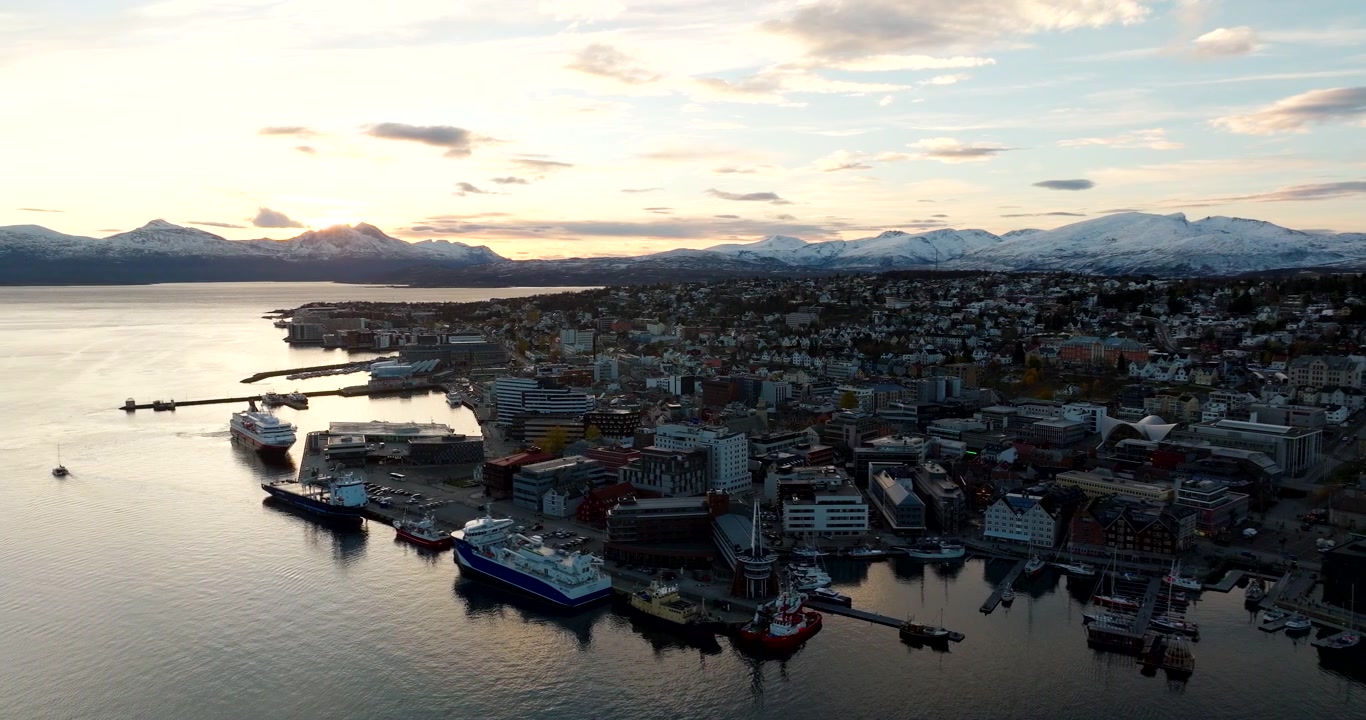 Luftaufnahme, Hafen und Docks einer Küstenstadt mit Schiffen, mit schneebedeckten Bergen im Hintergrund bei frühem Winterlicht in Tromsø, Norwegen.