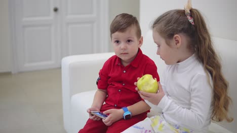 little pretty boy with mobile phone sitting near a little girl with green apple on the sofa and talking. shot in 4k