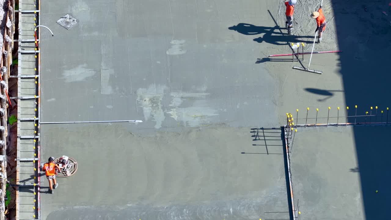 Two workers in safety gear level wet concrete with tools at a sunlit construction site