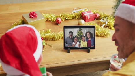 padre y hijo caucásicos con sombreros de santa usando una tableta para una videollamada de navidad con la familia en la pantalla