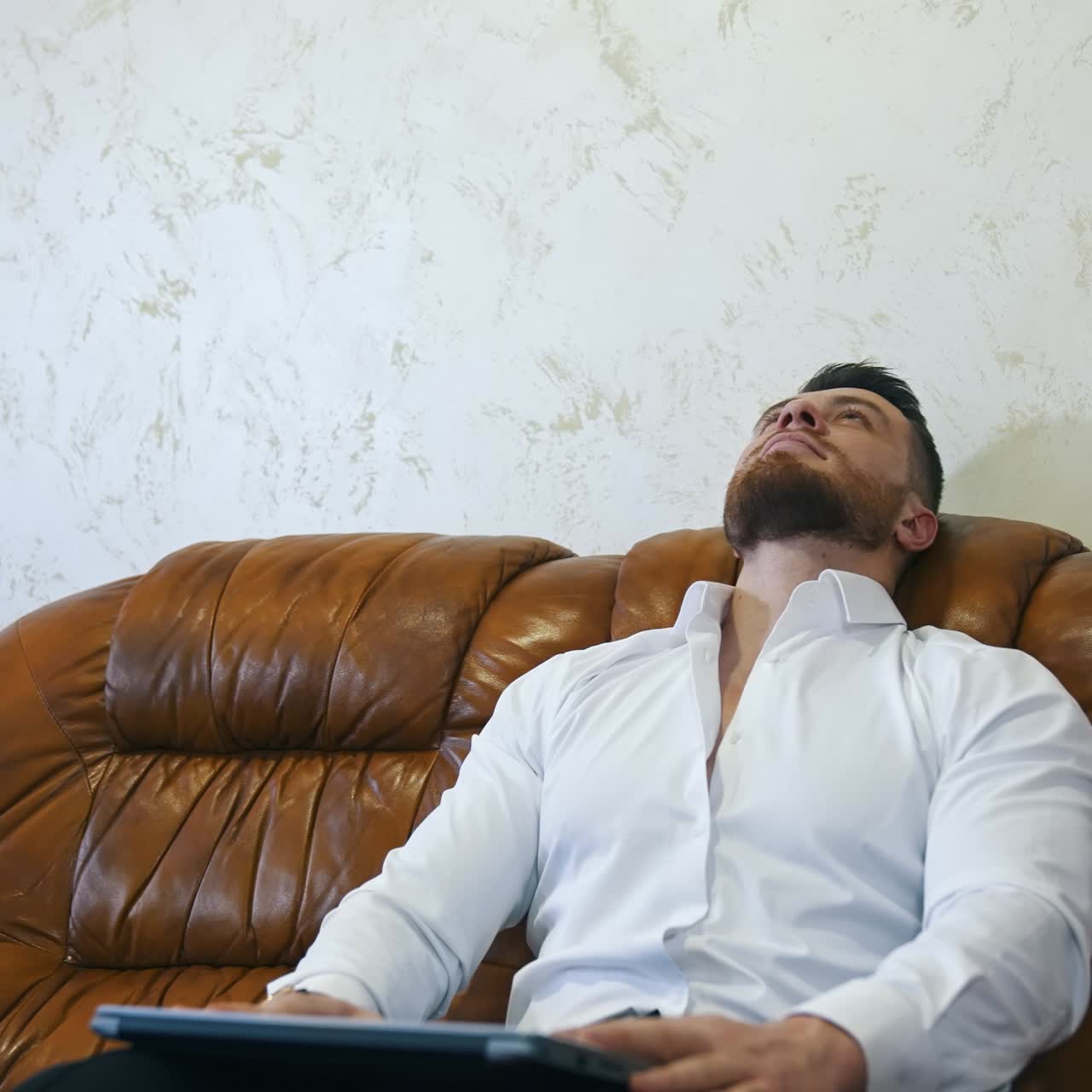 Freelancer using laptop while sitting on sofa. Tired businessman works online in business trip. Man resting on couch in hotel room. Business relaxing concept