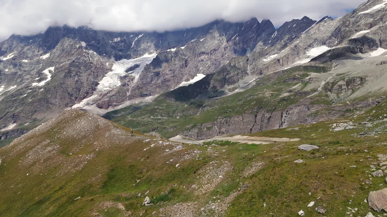 Aerial shot panning left to follow a man walking toward the summit of a sunny mountain in Cervinia, Italy