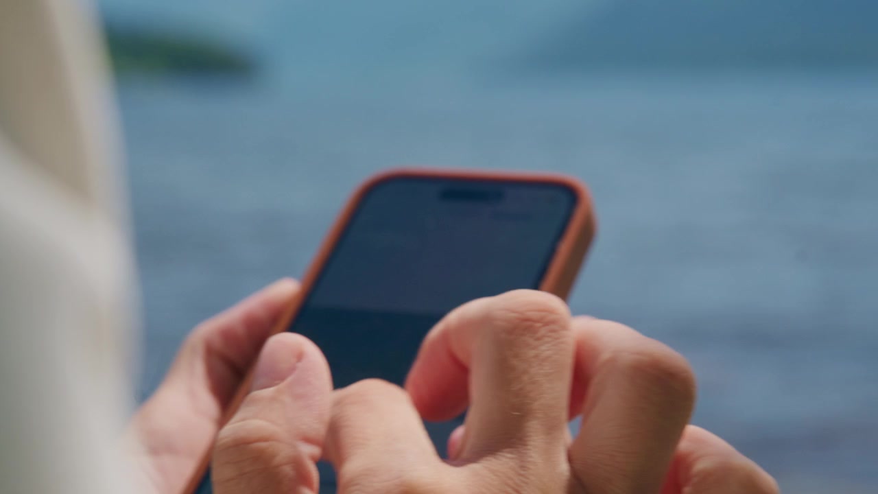 Primo piano di mani che digitano su uno smartphone vicino all'acqua. Le dita toccano lo schermo con concentrazione. Persona rilassata, che si gode il momento al mare. Ideale per social media, viaggi o temi di comunicazione.