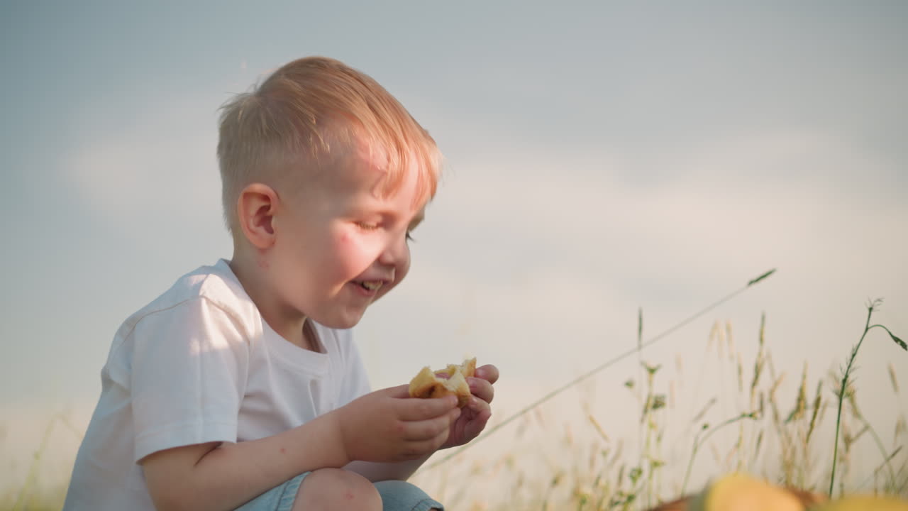 A woman in a blue dress joyfully takes a bite from her son's snack as they share a cheerful moment together in a grassy field, highlighting a tender, playful bond between mother and child