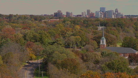 Vista-De-Una-Carretera-Y-Una-Iglesia-Entre-árboles-De-Otoño-Con-El-Horizonte-De-La-Ciudad-En-El-Horizonte