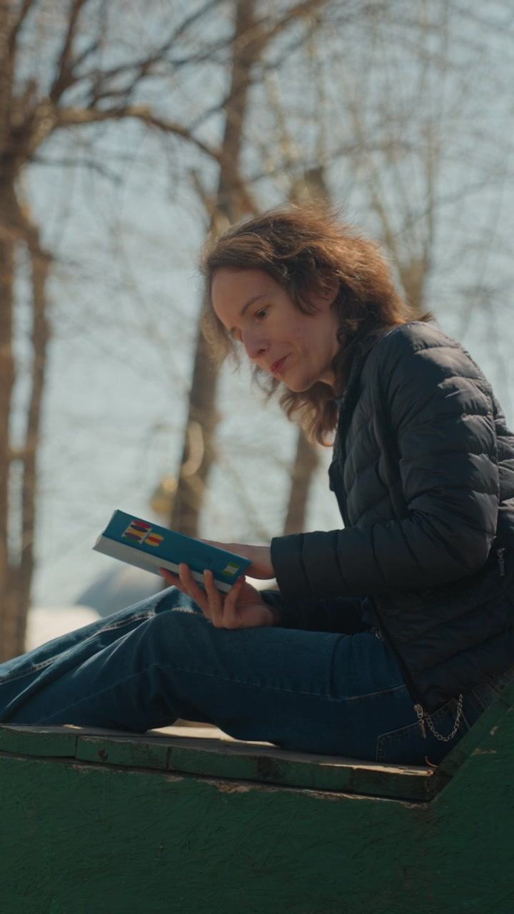 Close up of woman reading book outdoors on green bench with bare trees in background. Woman holds blue hardcover and appears engaged wearing dark jacket and jeans. Soft natural light creates