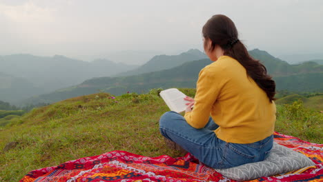 brown haired woman reading a book on a hill with mountain view