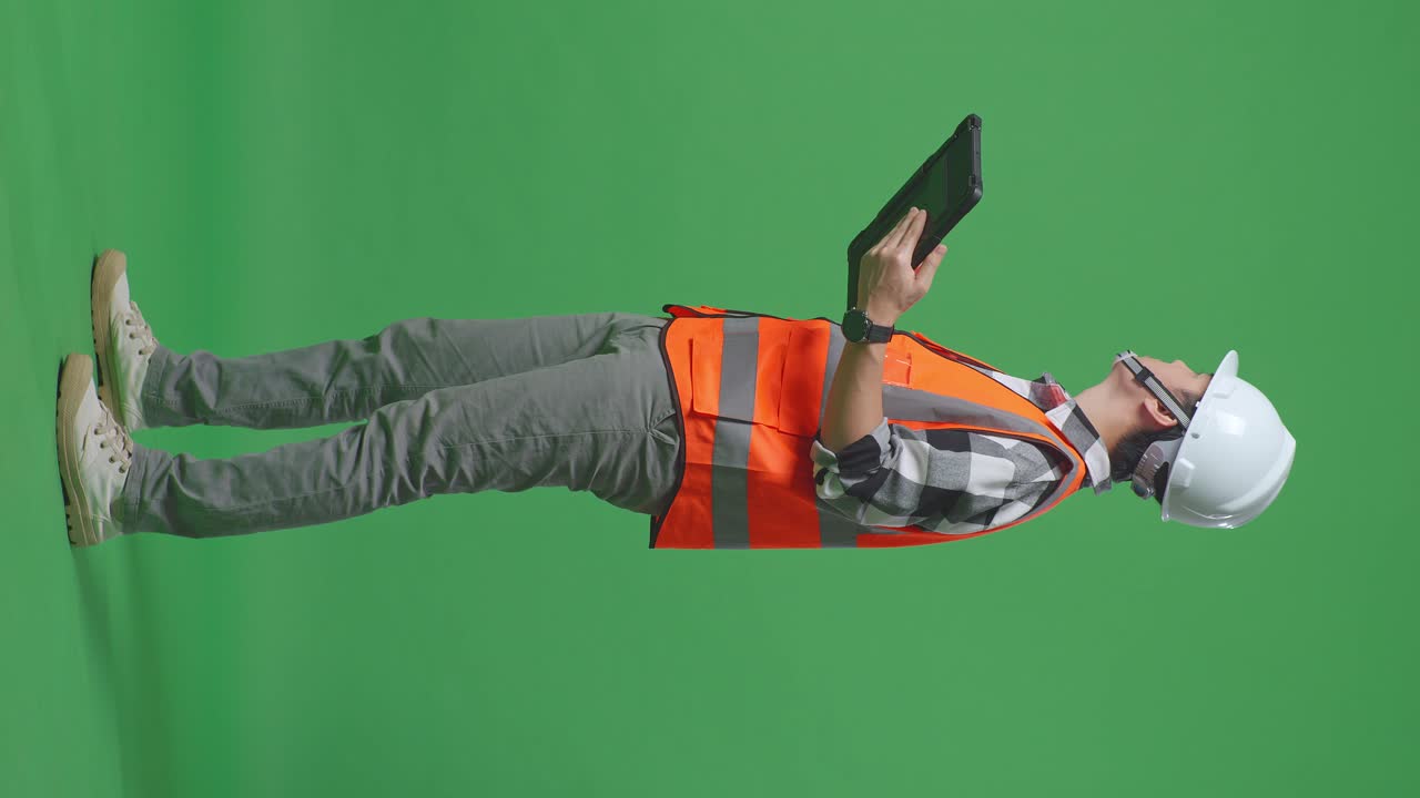 Full Body Side View Of Asian Male Engineer With Safety Helmet Looking At The Tablet In His Hand And Looking Around While Standing In The Green Screen Background Studio
