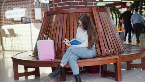 young lady seated on mall bench engrossed in book, two men walking in the background, leg crossed, casual attire, peaceful shopping environment