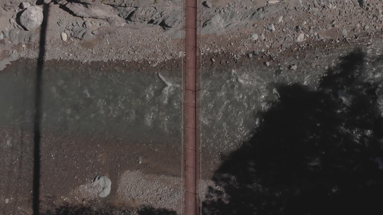 dos personas caminando a través de un puente colgante de cuerda de alambre de metal sobre el agua del río rocoso que fluye por el valle de la montaña kabayan benguet filipinas de arriba hacia abajo vista gran angular de ojo de pájaro