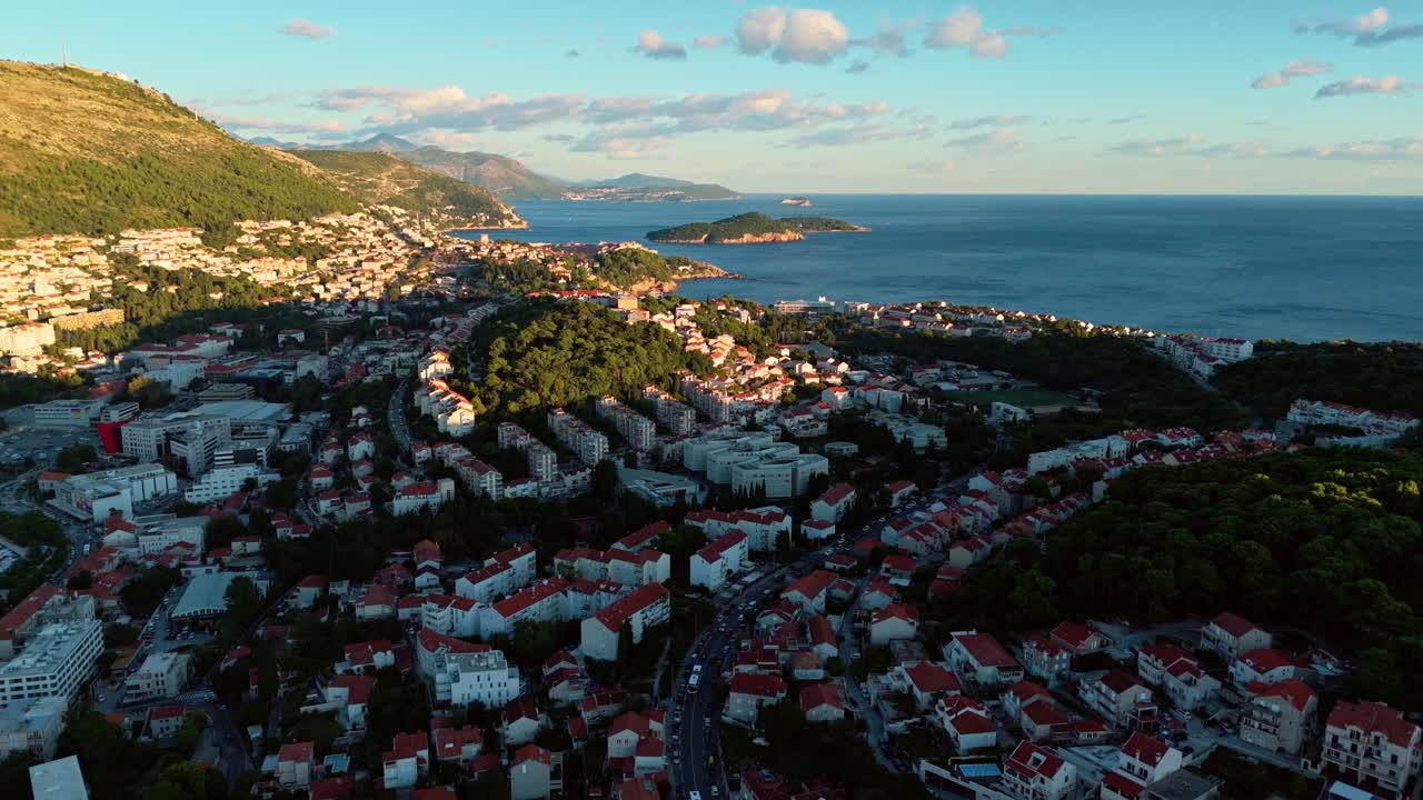 Aerial high angle drone shot over Dubrovnik during golden hour sunset, capturing the city’s red rooftops, surrounding mountains, and the calm Adriatic Sea stretching to the glowing ocean horizon