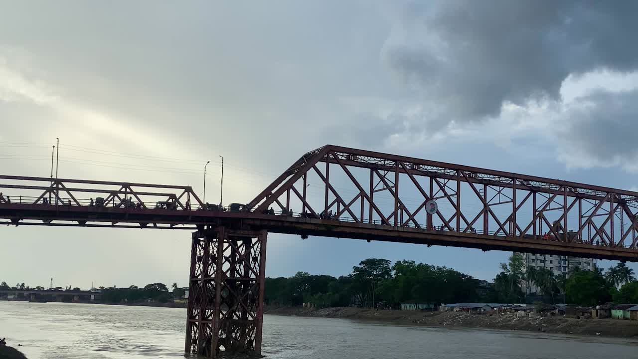 극적인 하늘을 배경으로 방글라데시 강 위의 금속 트러스 다리에서 통근하는 보행자와 차량