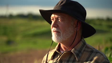 Close-up-portrait-of-senior-caucasian-good-looking-wise-man-farmer-in-a-hat-looking-at-the-side,-turning-face-to-the-camera-in-a-field