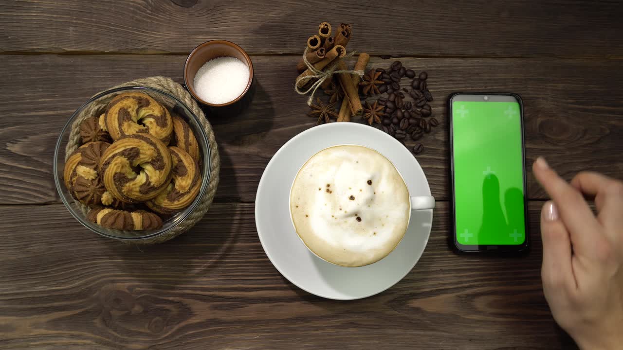 una mano femenina presiona la pantalla verde del teléfono inteligente tendido en la mesa con una taza de café.