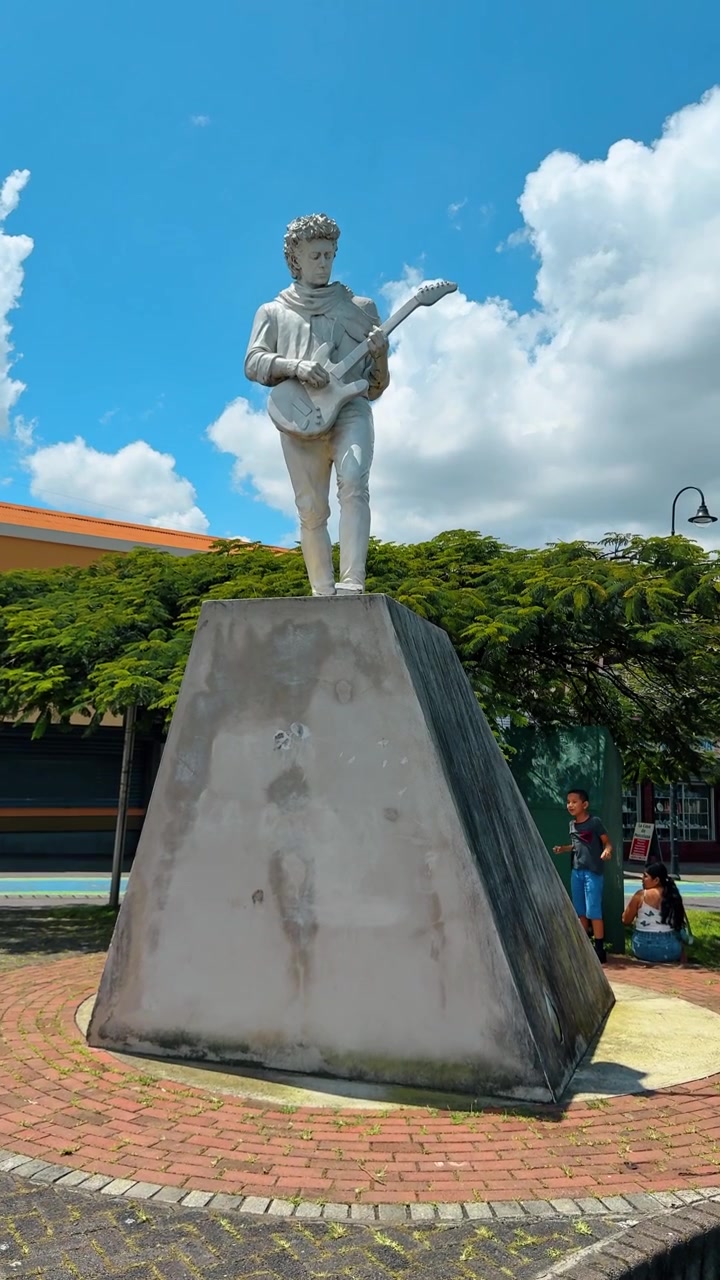 San Jose, Costa Rica, 1 agosto 2025: Statua di un chitarrista uomo in piedi su un piedistallo di cemento. Statua bianca che raffigura un musicista che suona una chitarra elettrica contro un cielo blu nuvoloso in Costa Rica.