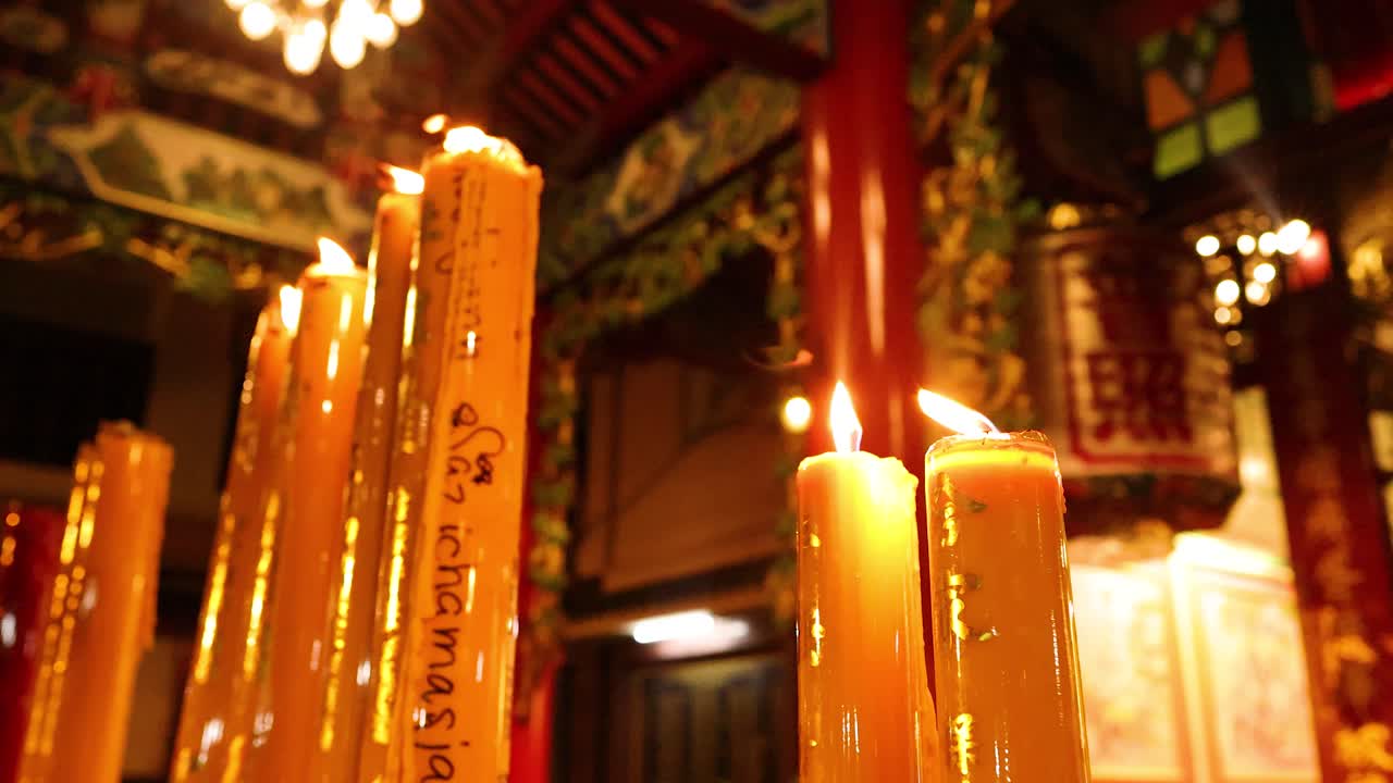 Tall candles burn steadily in richly decorated Asian temple, warm ambient lighting, slight camera movement