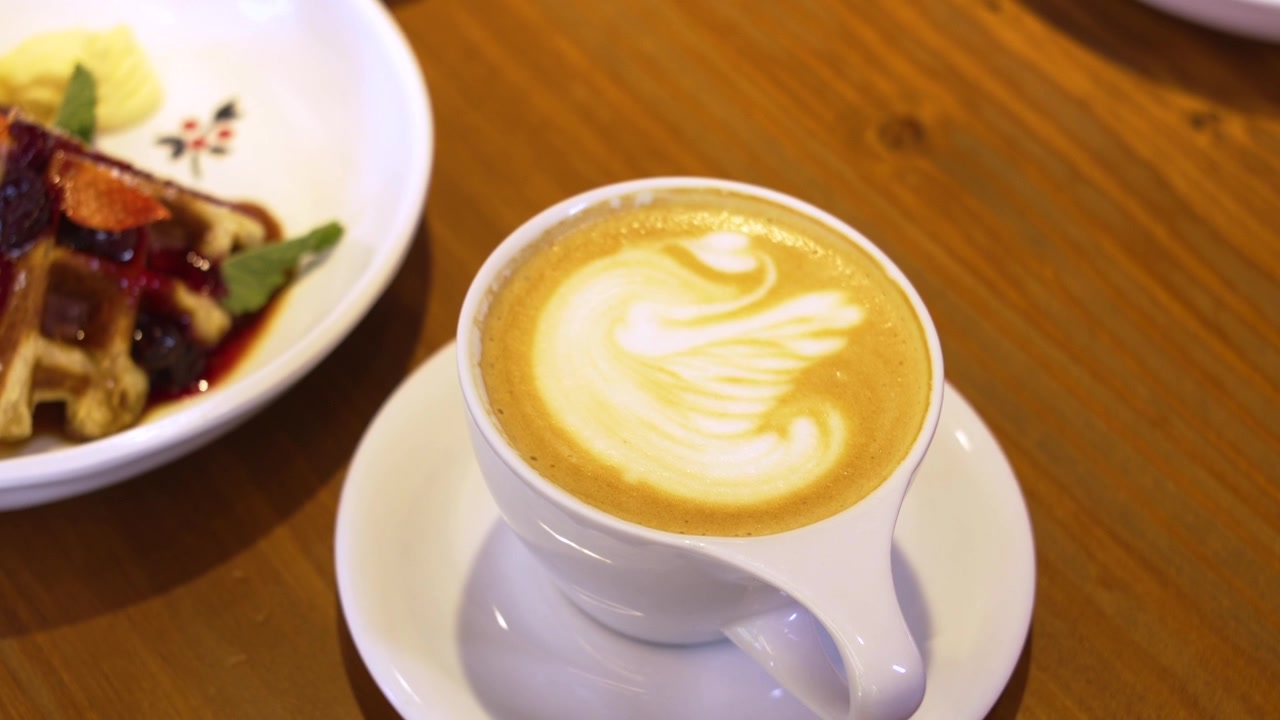 Latte Art en taza de cerámica blanca servido con postre de gofre gourmet en mesa de cafetería, café de especialidad.