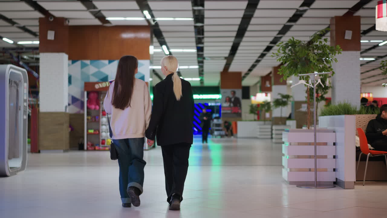 Rear view of two ladies walking hand in hand inside modern mall corridor, dressed in casual and formal outfits, creating warm moment of companionship