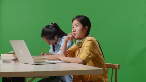 asian woman students thinking and looking around while sitting and writing into the notebooks on a table with a laptop in the green screen background classroom