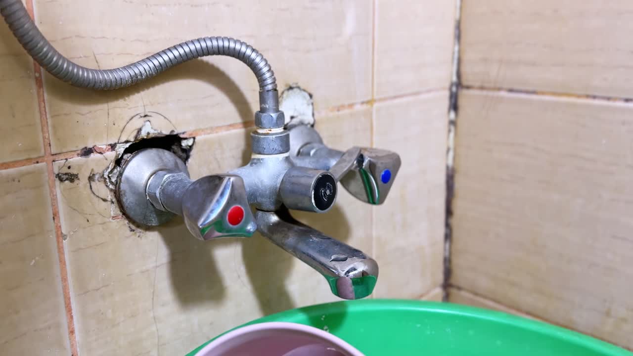 Primo piano di un rubinetto miscelatore a parete con manopole rosse e blu sopra un lavabo in plastica verde in un angolo di bagno piastrellato, che mostra l'impianto idraulico interno, l'attacco del tubo e lo stucco macchiato