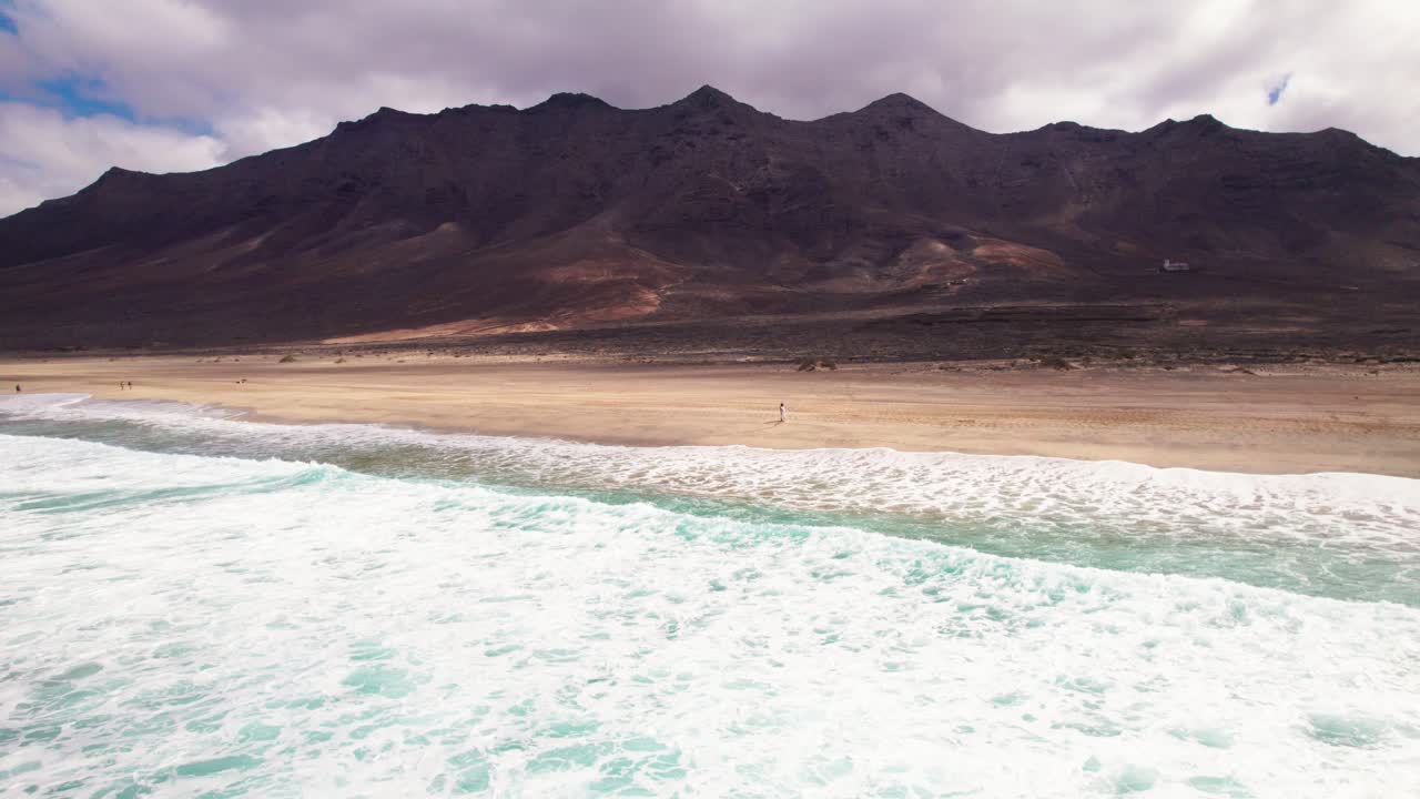 Aerial view from the ocean of woman in white dress on secluded Cofete Beach, Fuerteventura of turqoise waves crashing on the shoreline with endless golden sands and rugged mountains in the background