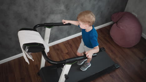aerial view of young boy standing on treadmill gripping handle with one hand and reaching for towel with other in relaxed indoor setting featuring wooden floor and casual fitness atmosphere