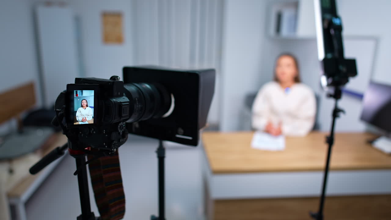 Camera on tripod filming a woman sitting at desk. Influencer creates a blog content in a studio. Blurred backdrop.