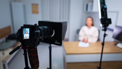 camera on tripod filming a woman sitting at desk. influencer creates a blog content in a studio. blurred backdrop.