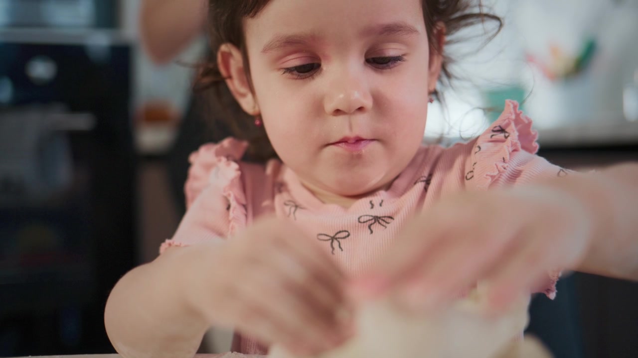Menina amassa massa com atenção concentrada. Ela veste um top rosa com babados, olhos fixos na tarefa. Mãos movem-se suavemente, moldando a mistura branca e macia. A luz quente da cozinha realça sua concentração