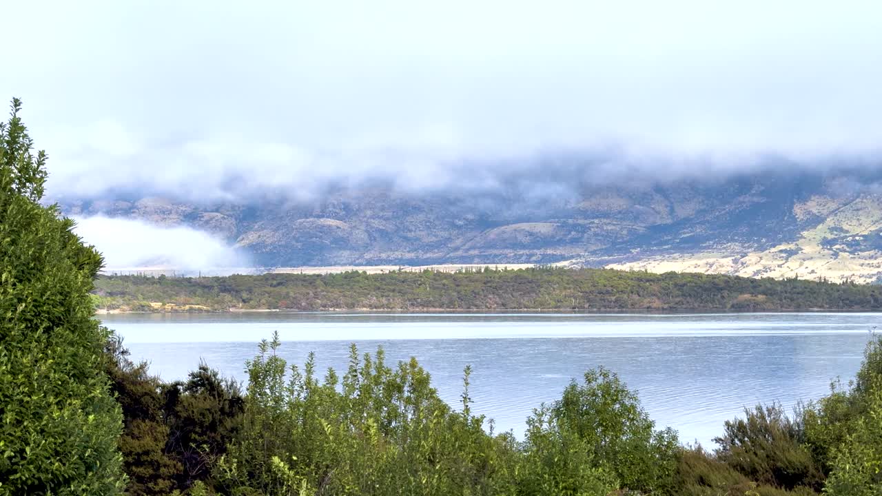 A tranquil view of Lake Wakatipu with mist-covered mountains and lush greenery under soft daylight in Queenstown, New Zealand