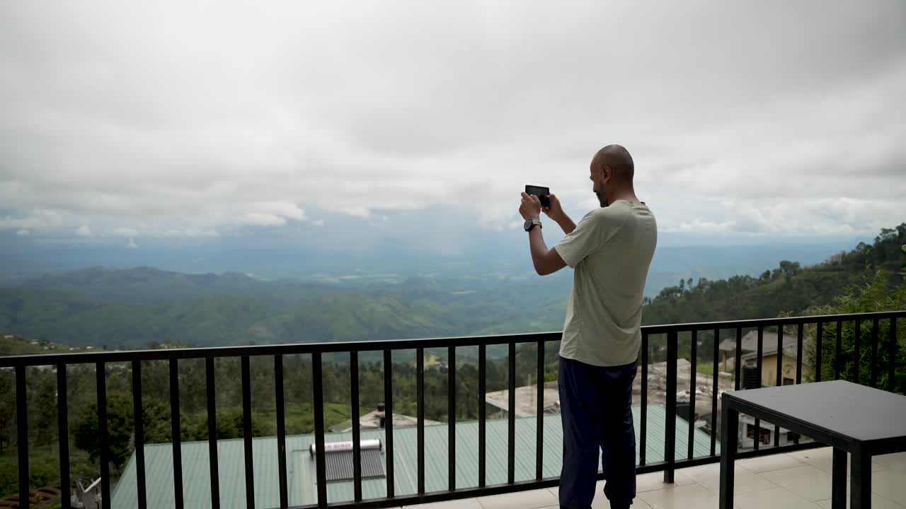 A man photographs a picturesque valley scenery using a smartphone while standing on a balcony.