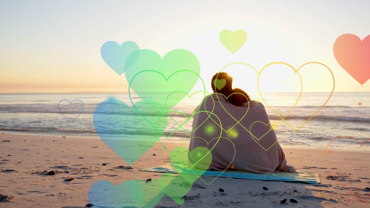Paar sitzt eingehüllt auf Matte mit Blick aufs Meer, tiefe Sonne lässt Herzen durchs Bild schweben für Marketingzwecke.